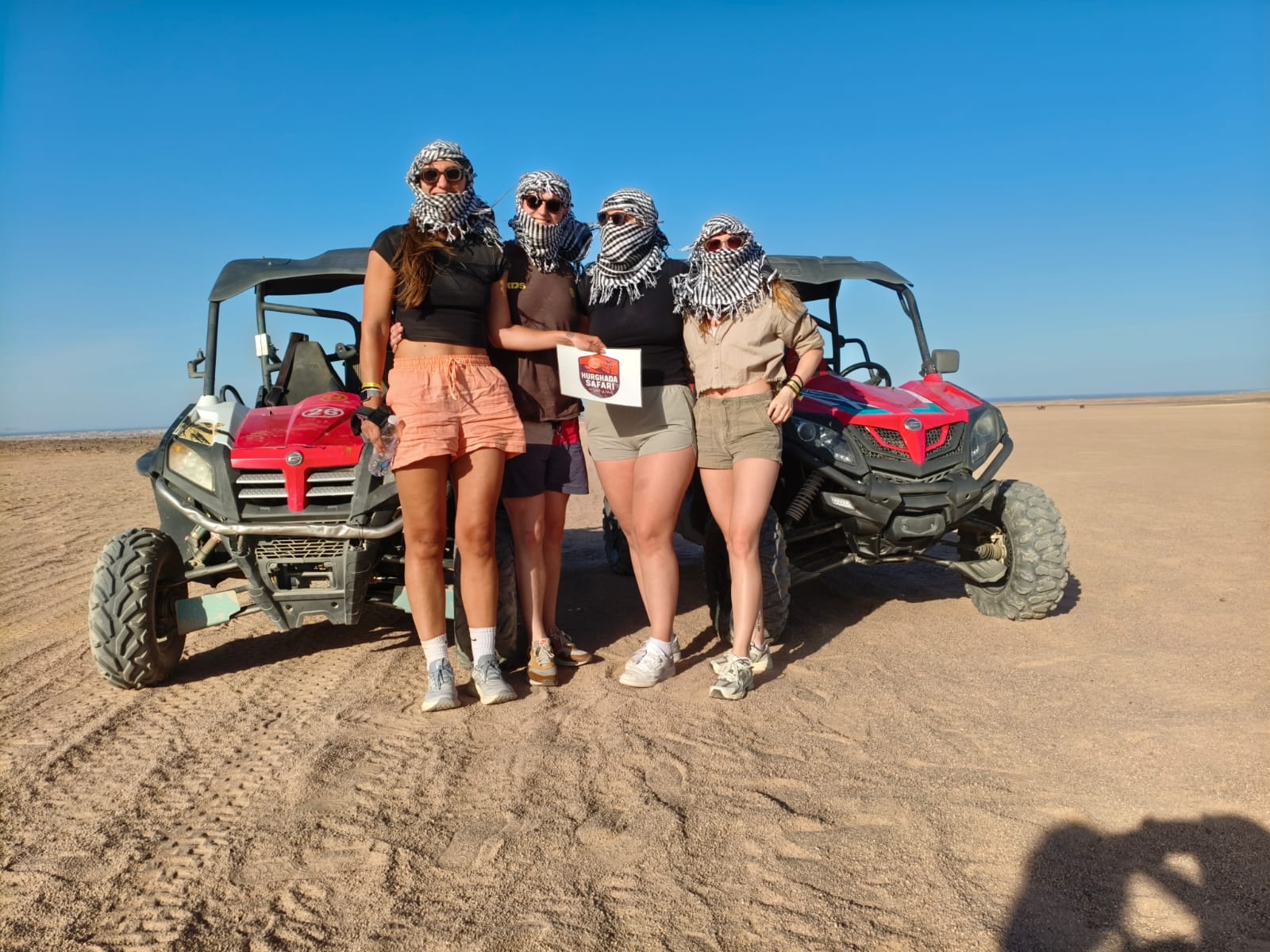 Group of friends with desert scarves holding the Hurghada Safari sign beside buggies in the desert